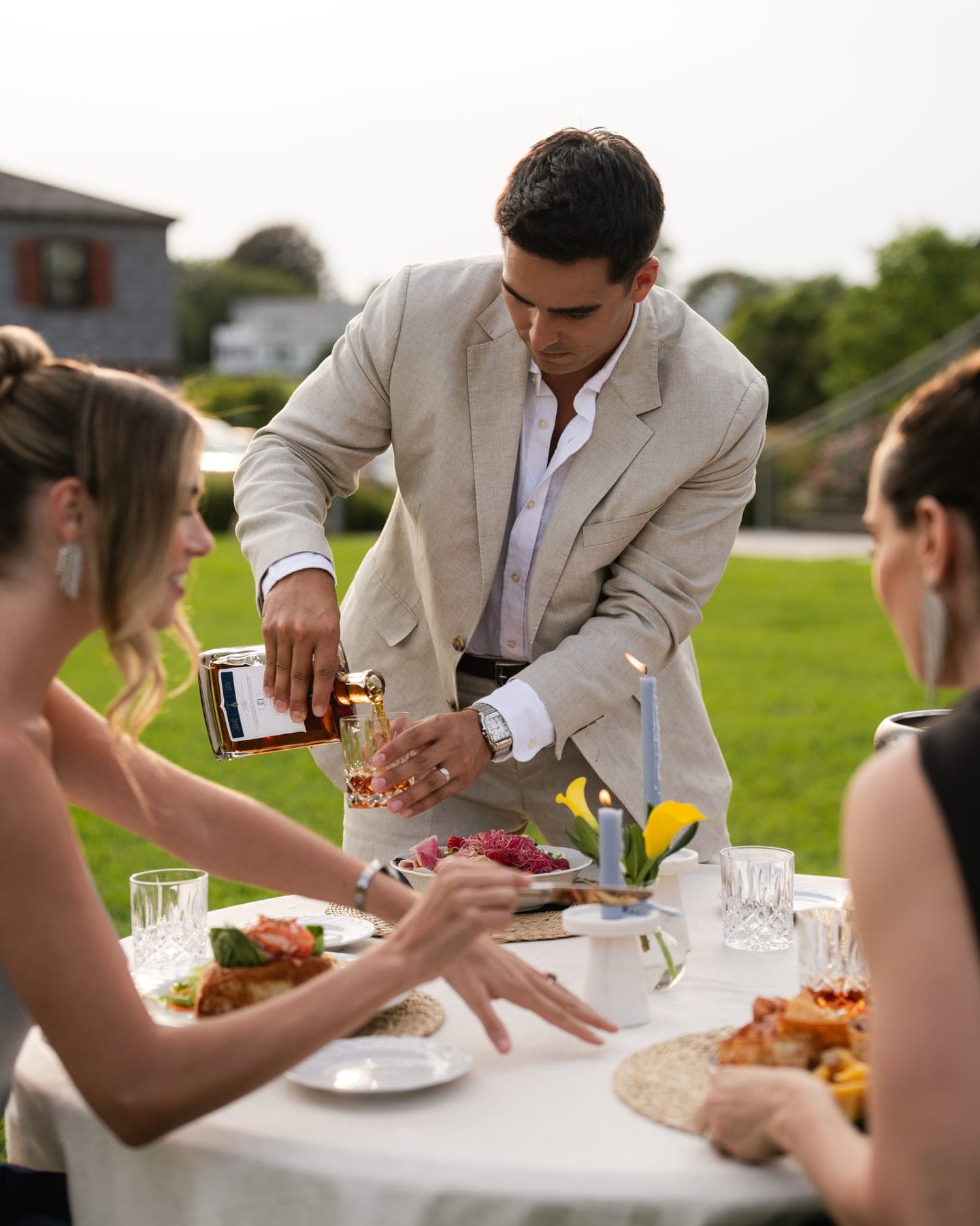 A host pouring bourbon for guests at an outdoor dinner table